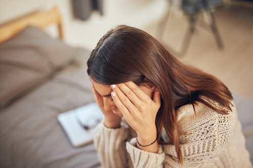 Attractive caucasian brunette dressed in beige sweater sitting on bed in bedroom and having headache.