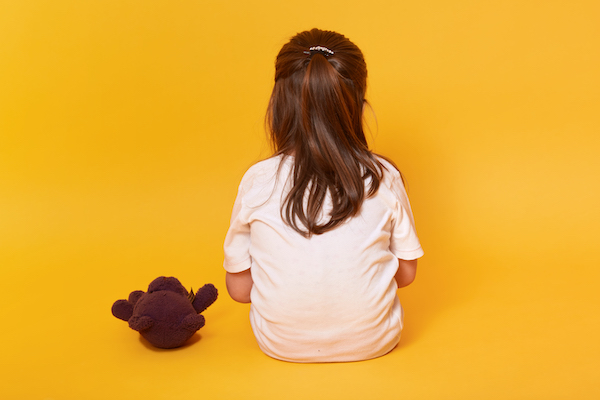 Little girl sitting backwards with brown teddy bear, toddler wearing white t shirt offended by someone, being in bad mood, feels sad and upset, posing isolated over yellow background. Emotions concept